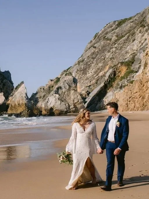 A couple in wedding attire walking hand in hand on a sandy beach with large rock formations in the background.