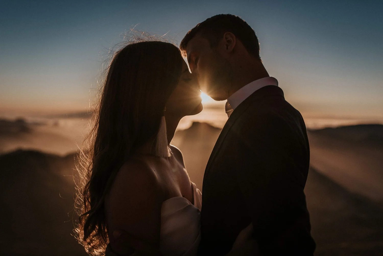 A couple stands close together on the beach during sunset, with their foreheads touching and the sun shining behind them.