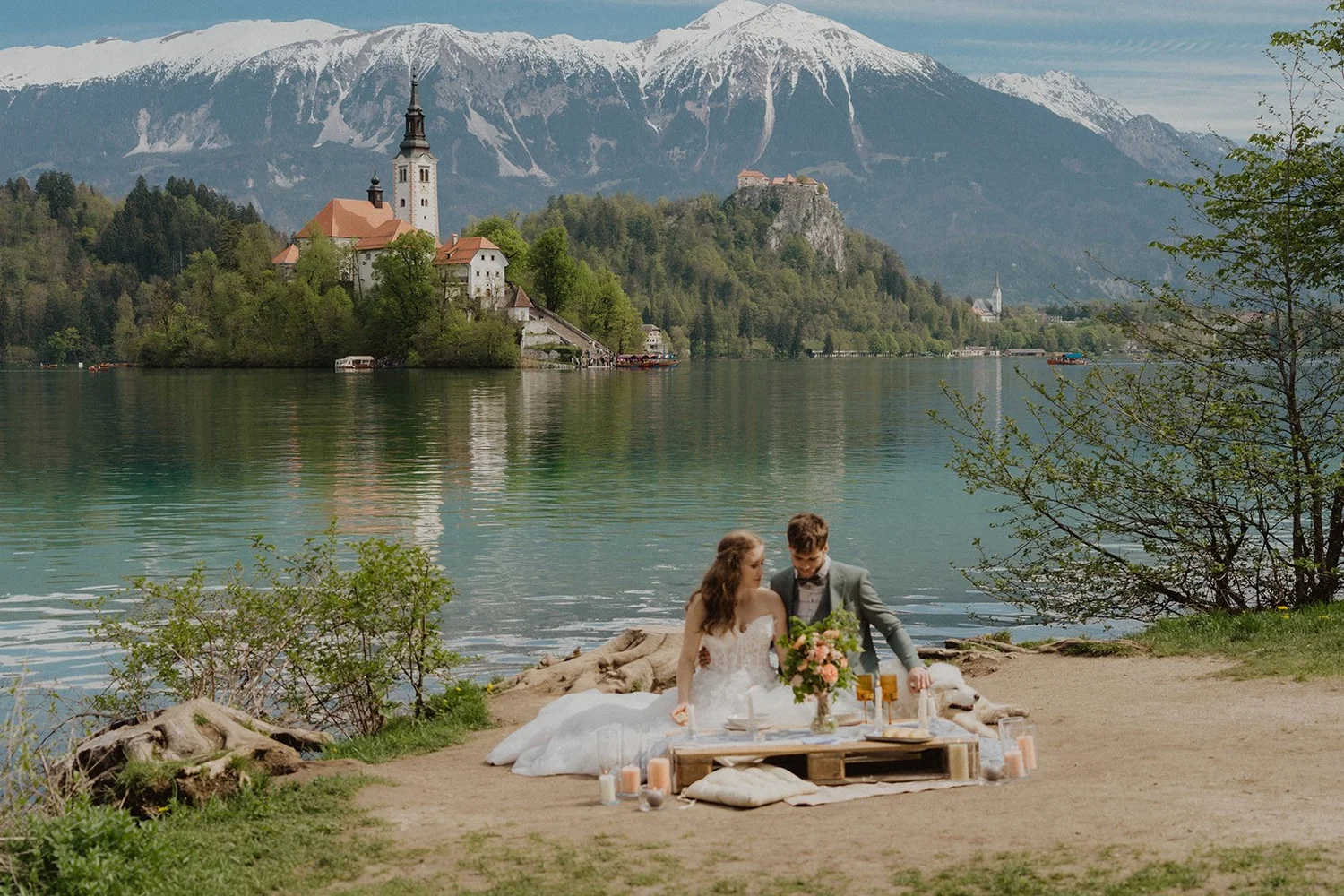 A bride and groom having a wedding celebration outdoors by a lake with mountains in the background. The bride is wearing a white wedding dress and the groom is in a gray suit. They are sitting on the ground next to a low table decorated with flowers, candles, and drinks on a blanket.