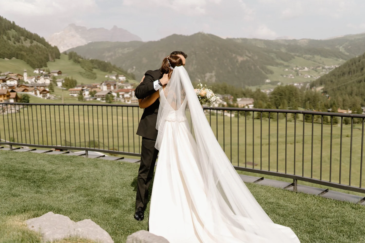 A bride and groom share a kiss on a balcony overlooking a picturesque mountain village and lush green hills.