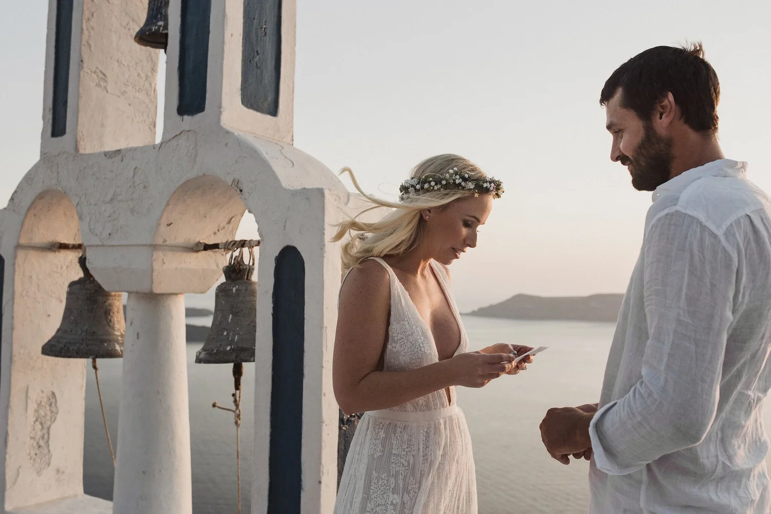 A couple is standing near a white Orthodox church with bells, exchanging rings during their wedding ceremony by the sea at sunset, with a mountain range in the background.