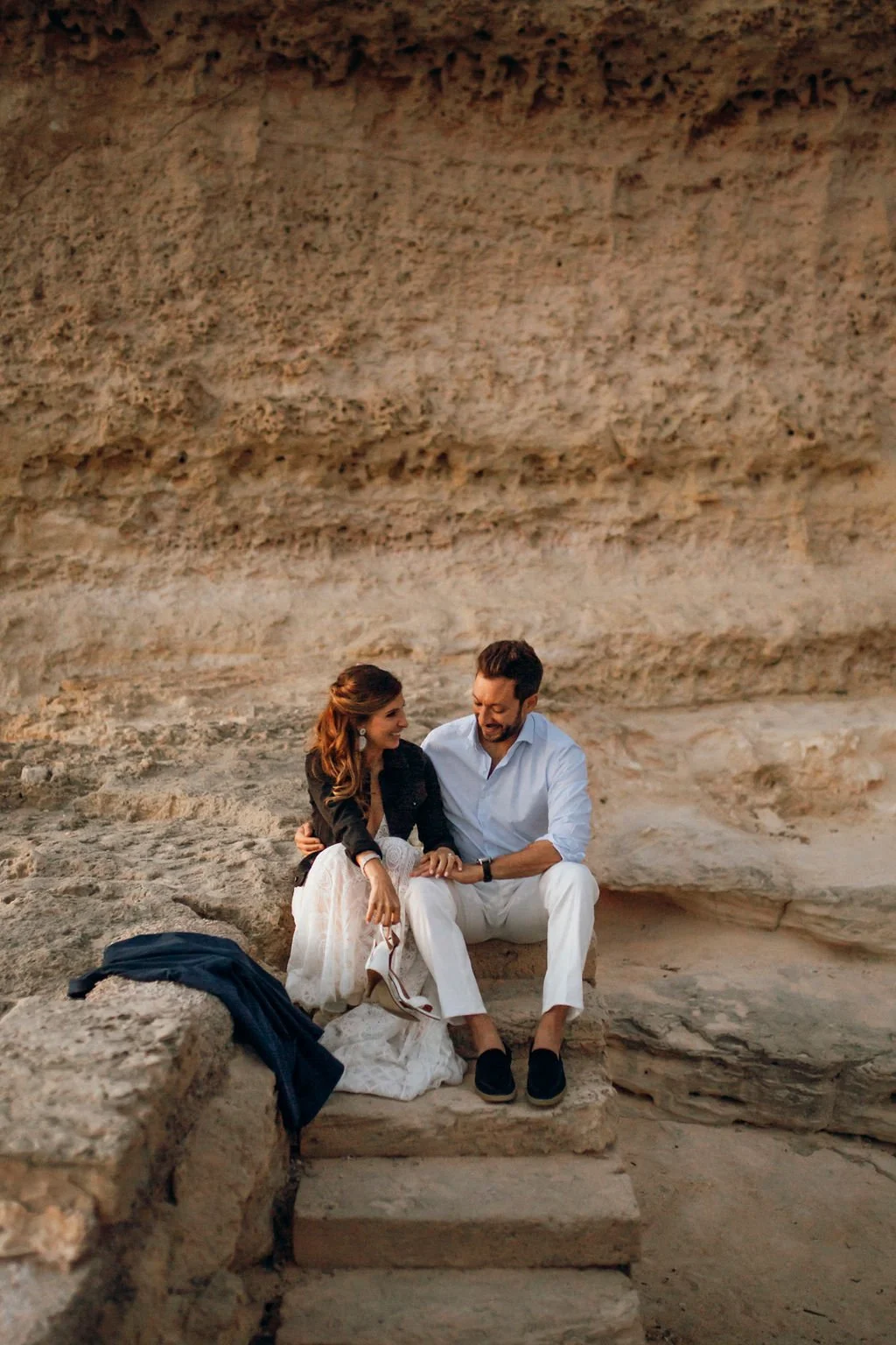 A couple sitting on stone steps inside a natural cave, smiling at each other, with a backpack and clothing nearby.