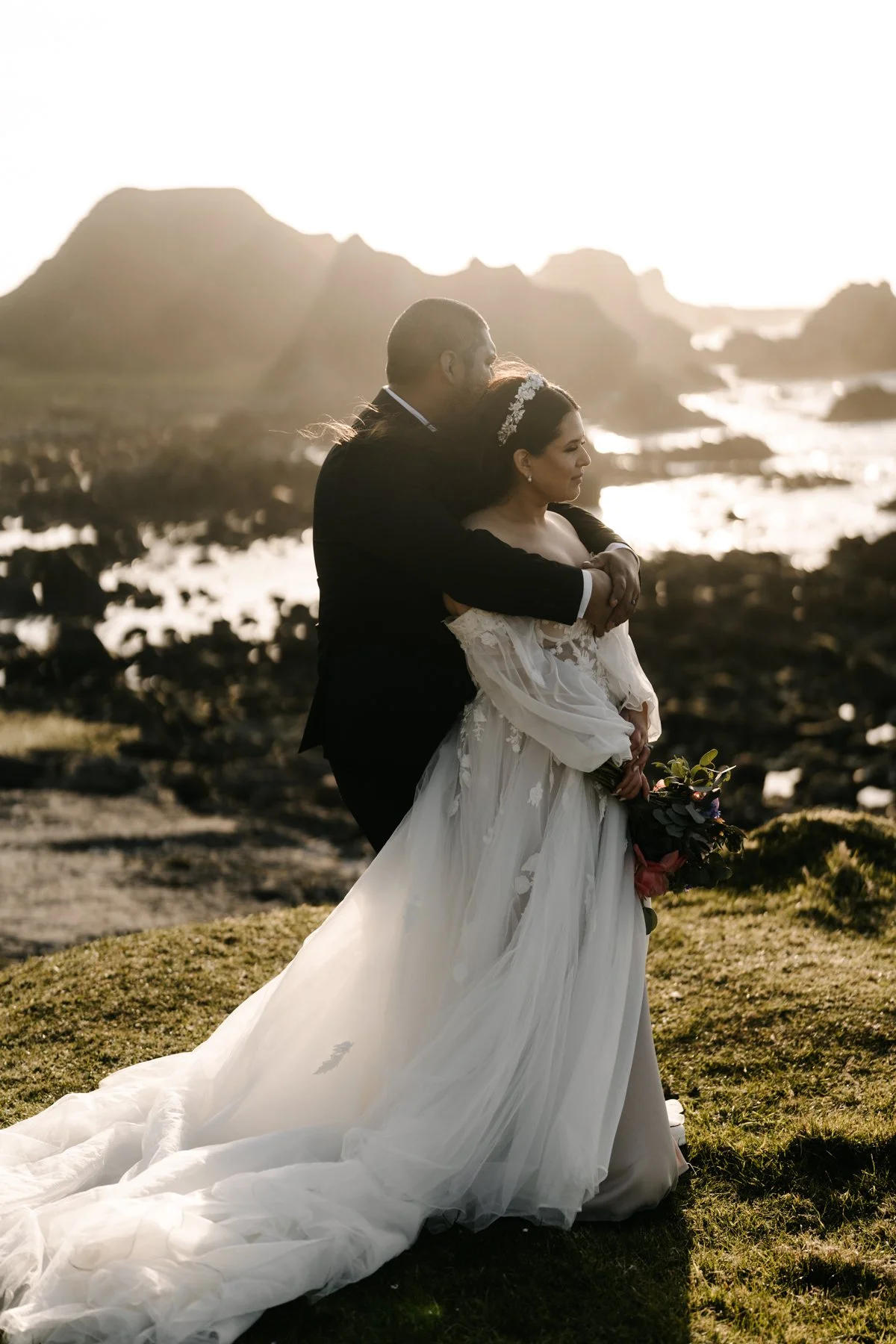 A bride and groom embracing outdoors at sunset with mountains and water in the background. The bride is in a white wedding gown holding a bouquet, and the groom is in a dark suit.