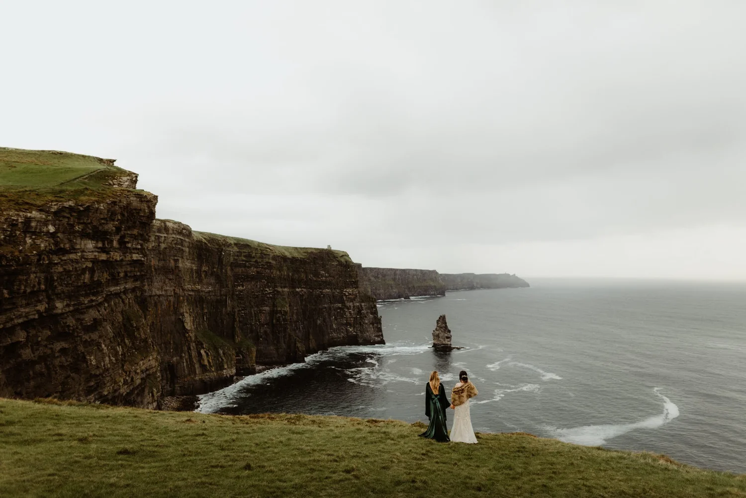 Two women standing on grass by the ocean, with cliffs and rock formations in the background on a cloudy day.