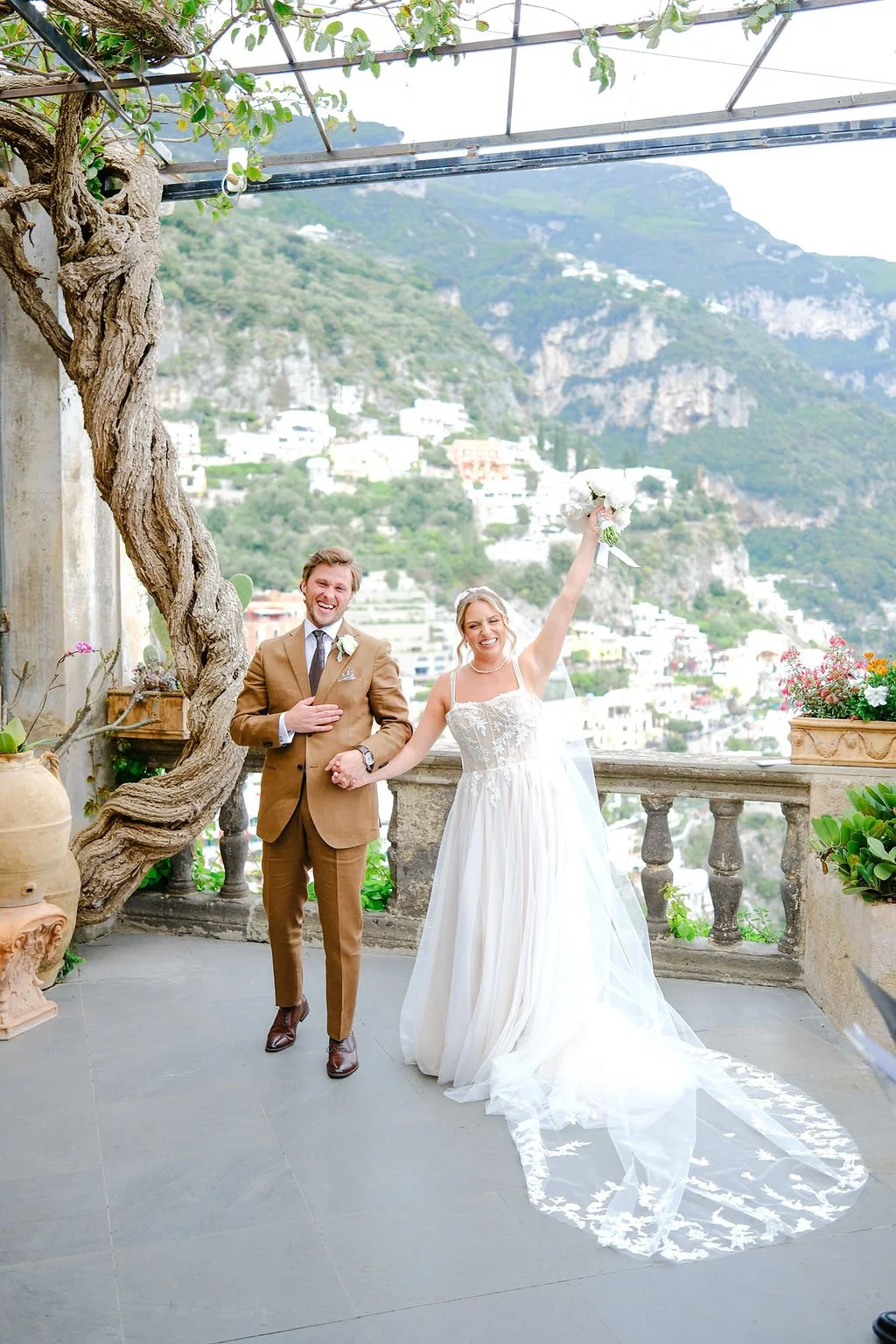 A newlywed couple stands on a terrace with a mountain view in the background. The bride in a white wedding dress is holding a bouquet and raising her arm enthusiastically. The groom in a brown suit is smiling and holding her hand. The setting include