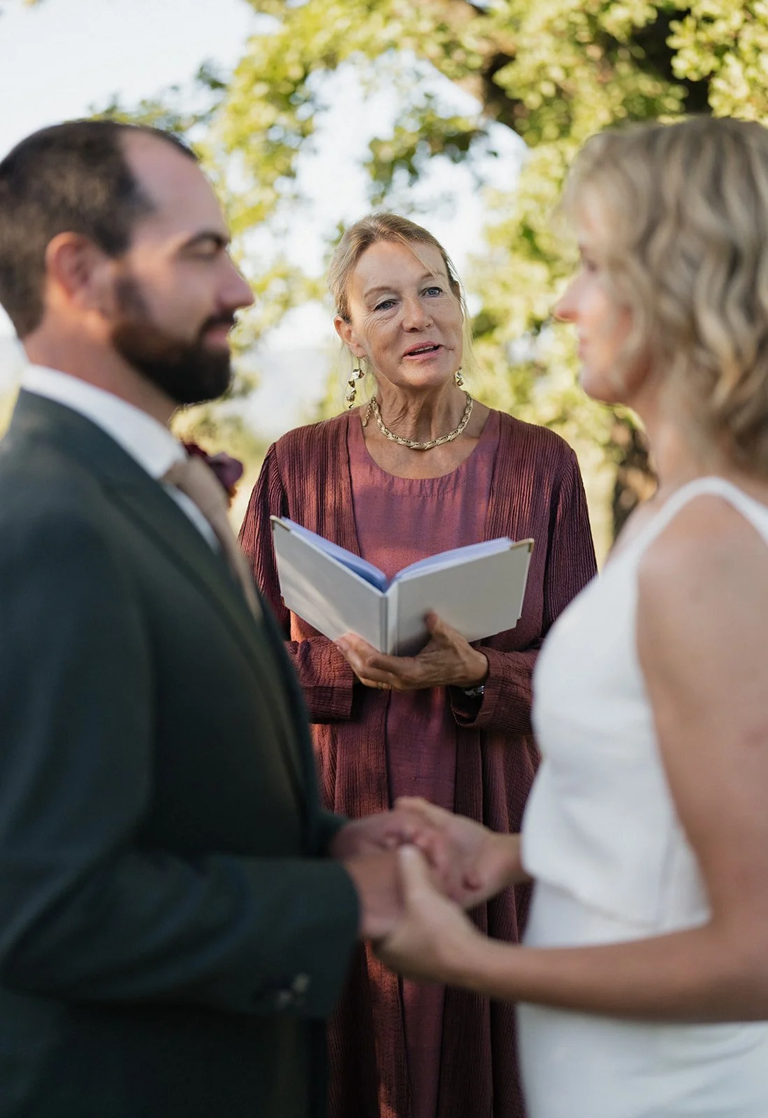 A couple getting married, holding hands and facing each other, with a officiant in the background reading from a book outdoors under a tree.
