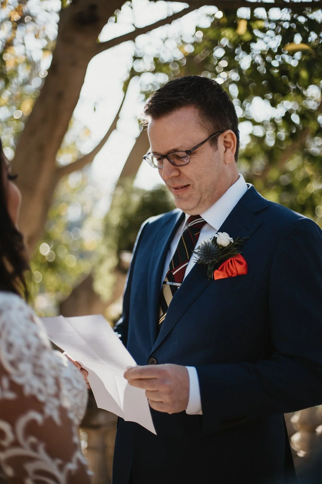 A groom in a blue suit and glasses is standing outdoors, exchanging vows with a bride in a lace dress, during a wedding ceremony under trees.