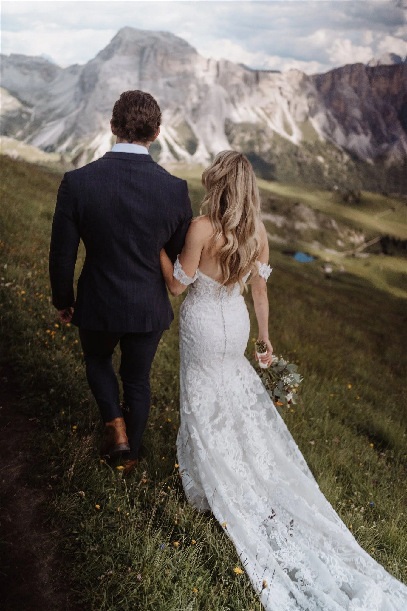 A couple in wedding attire walking through a grassy field with mountain peaks in the background.