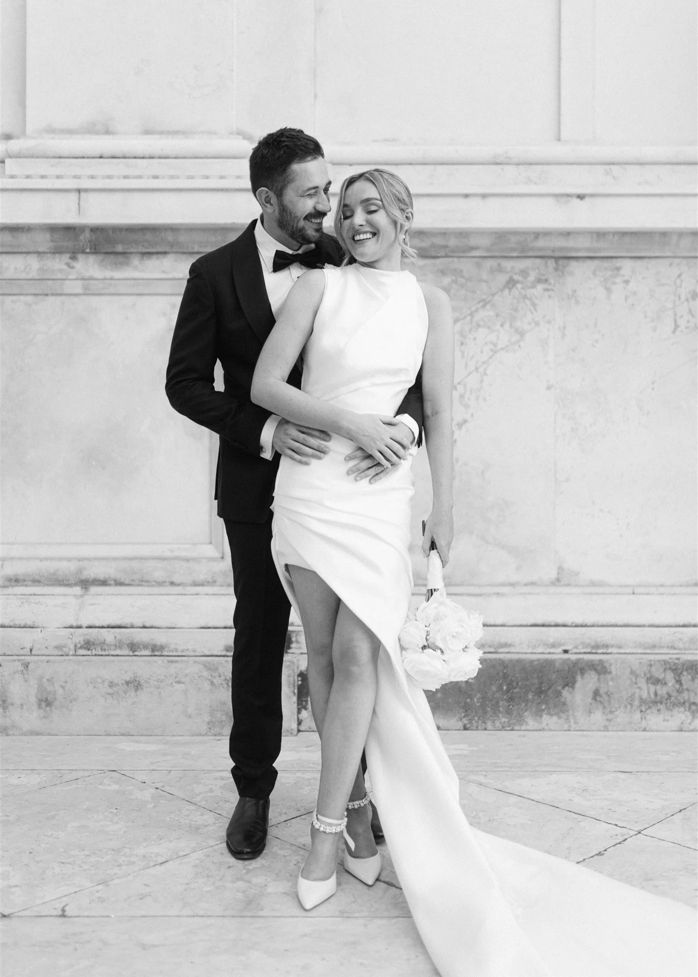 A black-and-white photo of a smiling bride and groom standing on steps. The groom wears a tuxedo and bow tie, and the bride wears a strapless wedding gown and heels, holding a bouquet of flowers.