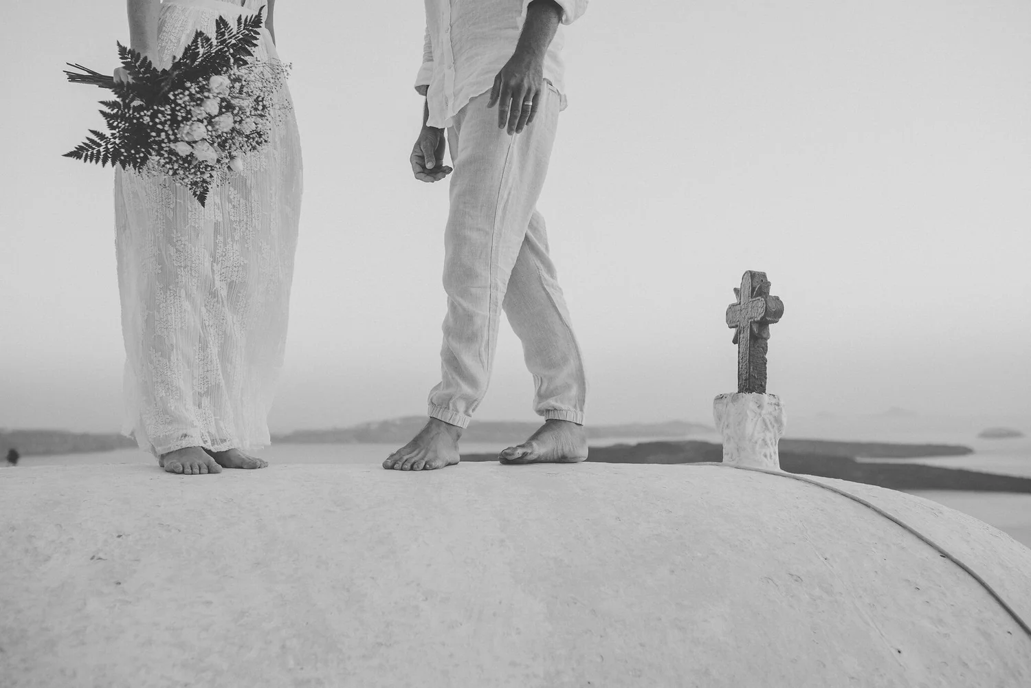 Black and white photo of two people standing barefoot on a flat surface, possibly on a beach or desert. One person is holding a bouquet of flowers. A small cross on a pedestal is visible near one person's feet.