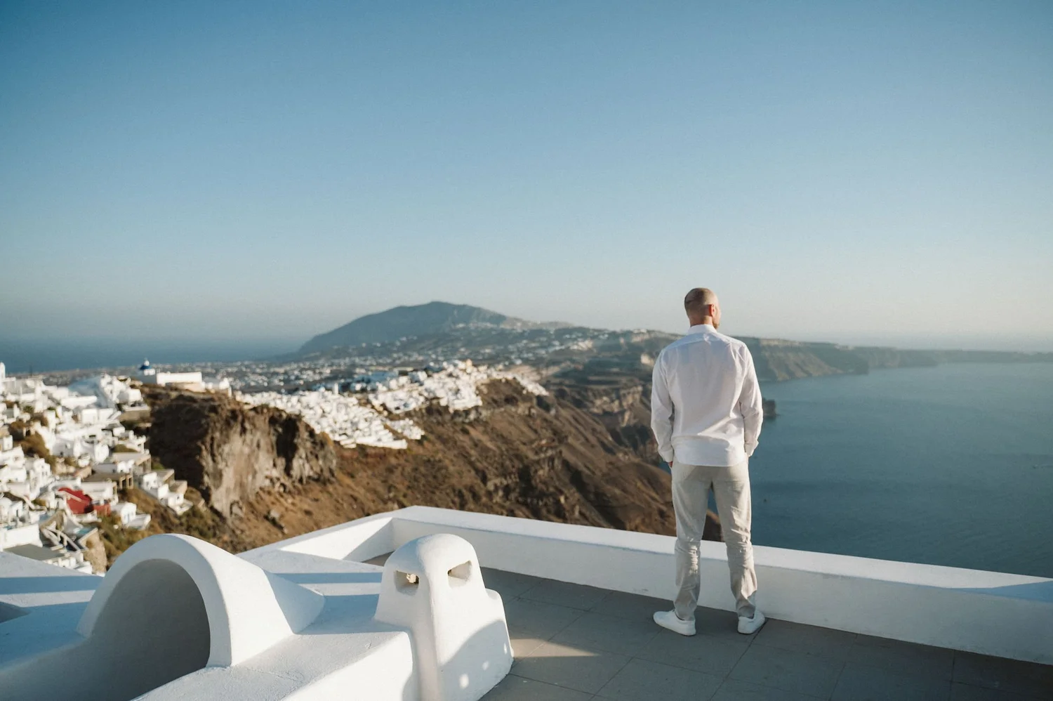 A man dressed in white stands on a white balcony overlooking a coastal town and sea, with a mountain in the background, under clear blue sky.