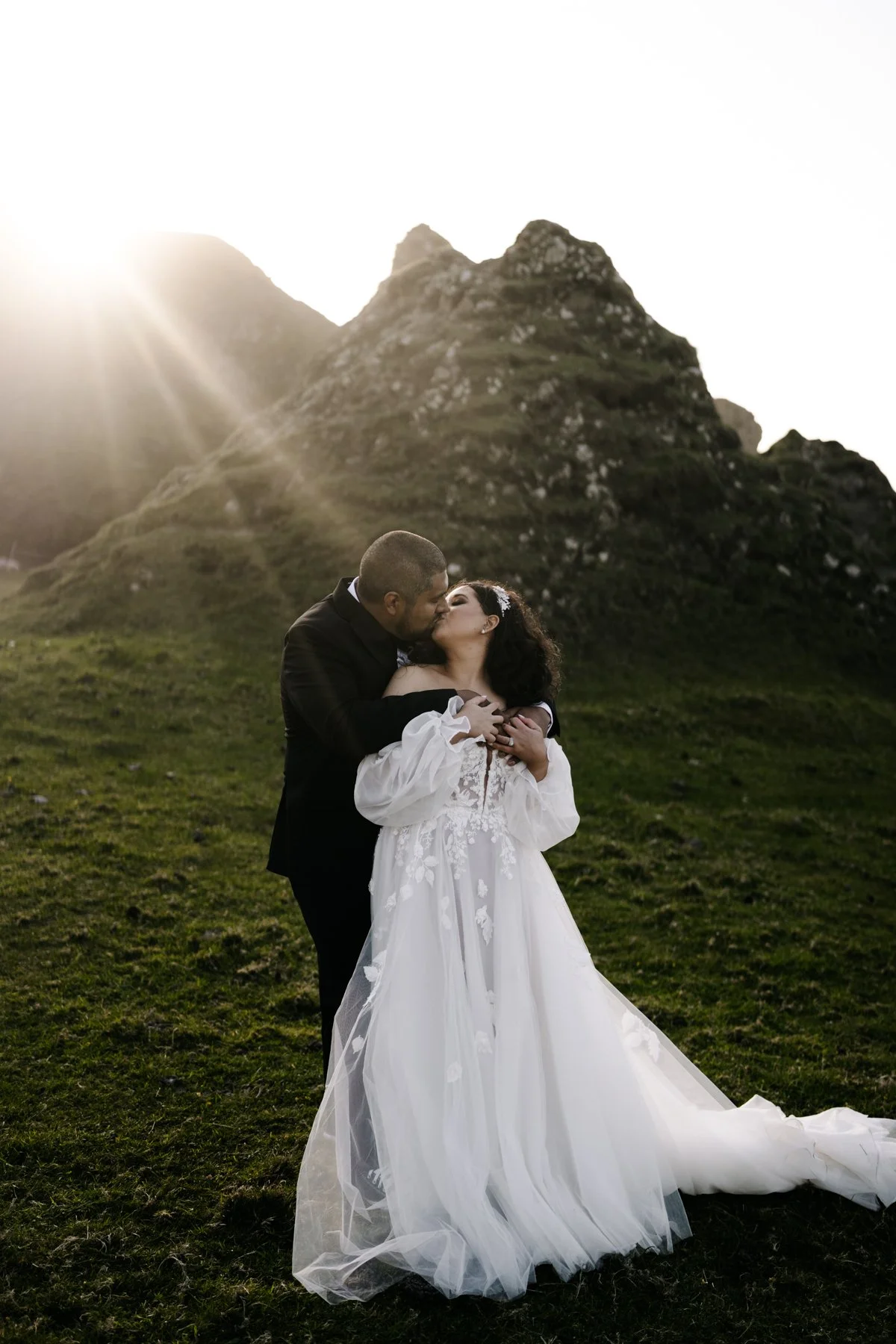 Elopement portrait of a bride and groom sharing a kiss outdoors at sunset with a mountain in the background. The bride wears a white wedding dress, and the groom wears a black suit.
