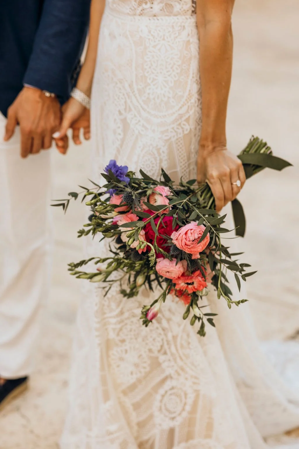 Bride holding a bouquet of pink, red, purple, and dark blue flowers, dressed in an intricate white lace wedding gown, with a smiling groom in a dark suit and white shirt holding her hand.