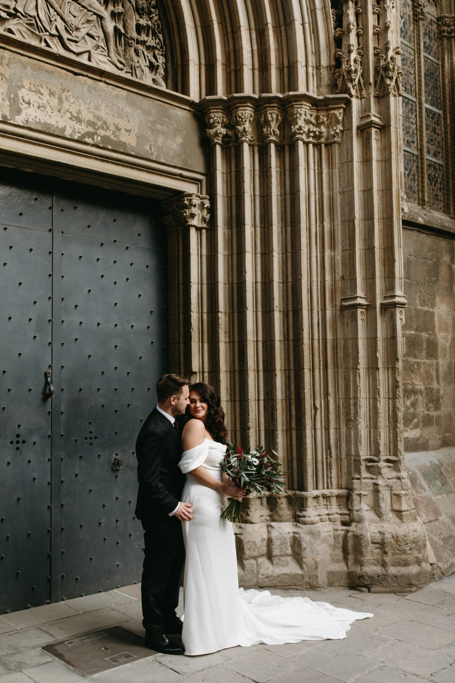 A couple dressed in wedding attire, standing outside a historic stone building with ornate architectural details, sharing a tender moment.