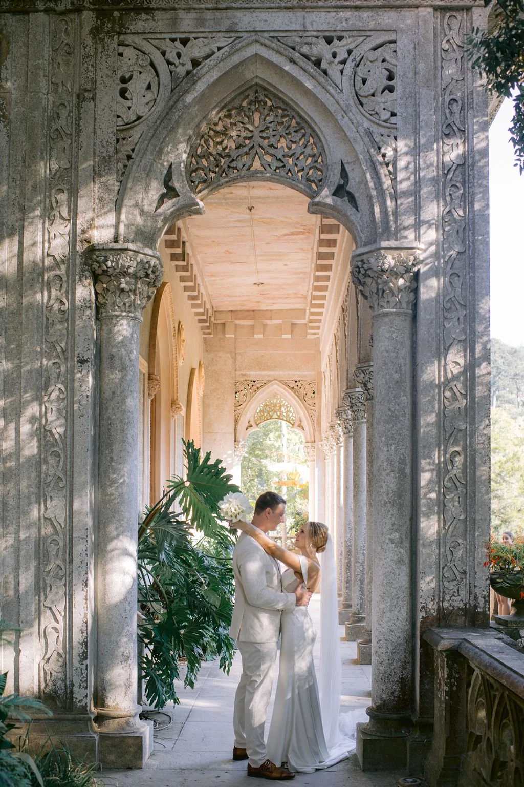A bride and groom standing under ornate stone archways, embracing each other, during their wedding ceremony outdoors surrounded by greenery and decorative architecture.