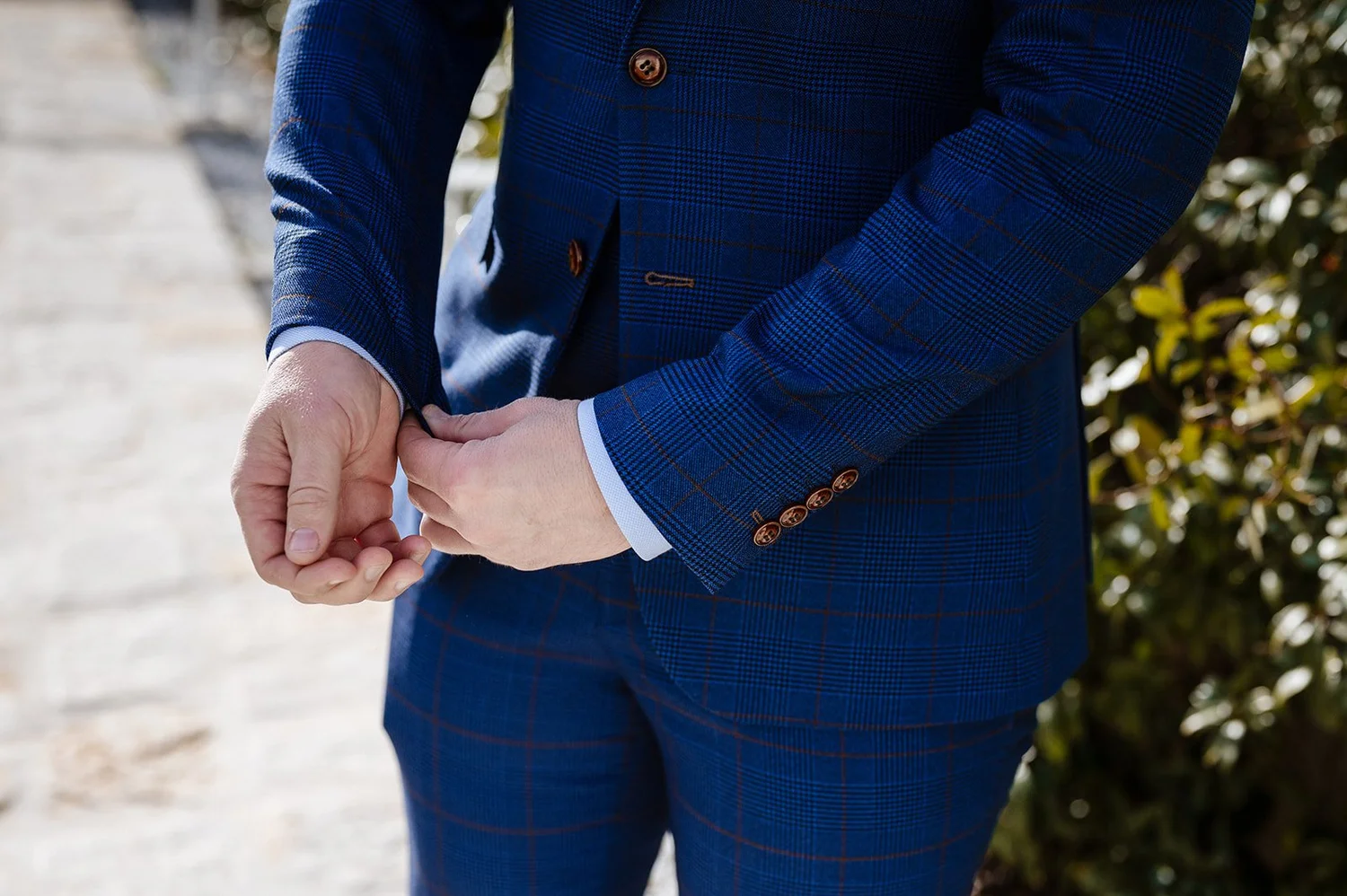 A person wearing a blue checkered suit with brown buttons, adjusting their cufflinks outdoors, with green foliage in the background.