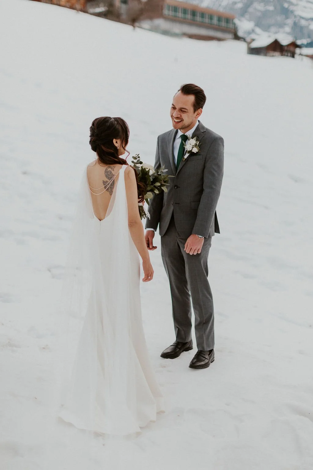 A bride and groom stand together outdoors on snow, smiling and looking at each other, with the bride holding a bouquet of flowers.