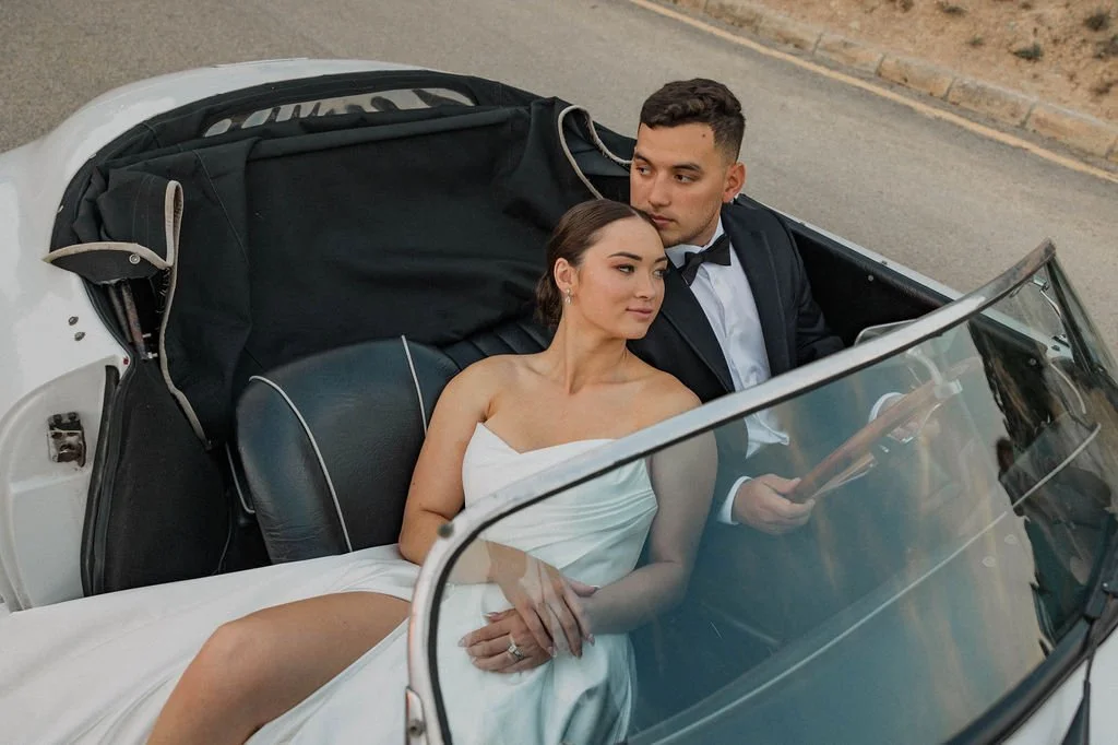 A bride and groom sitting in a vintage convertible car, with the bride in a white dress and the groom in a black tuxedo, on a street setting.