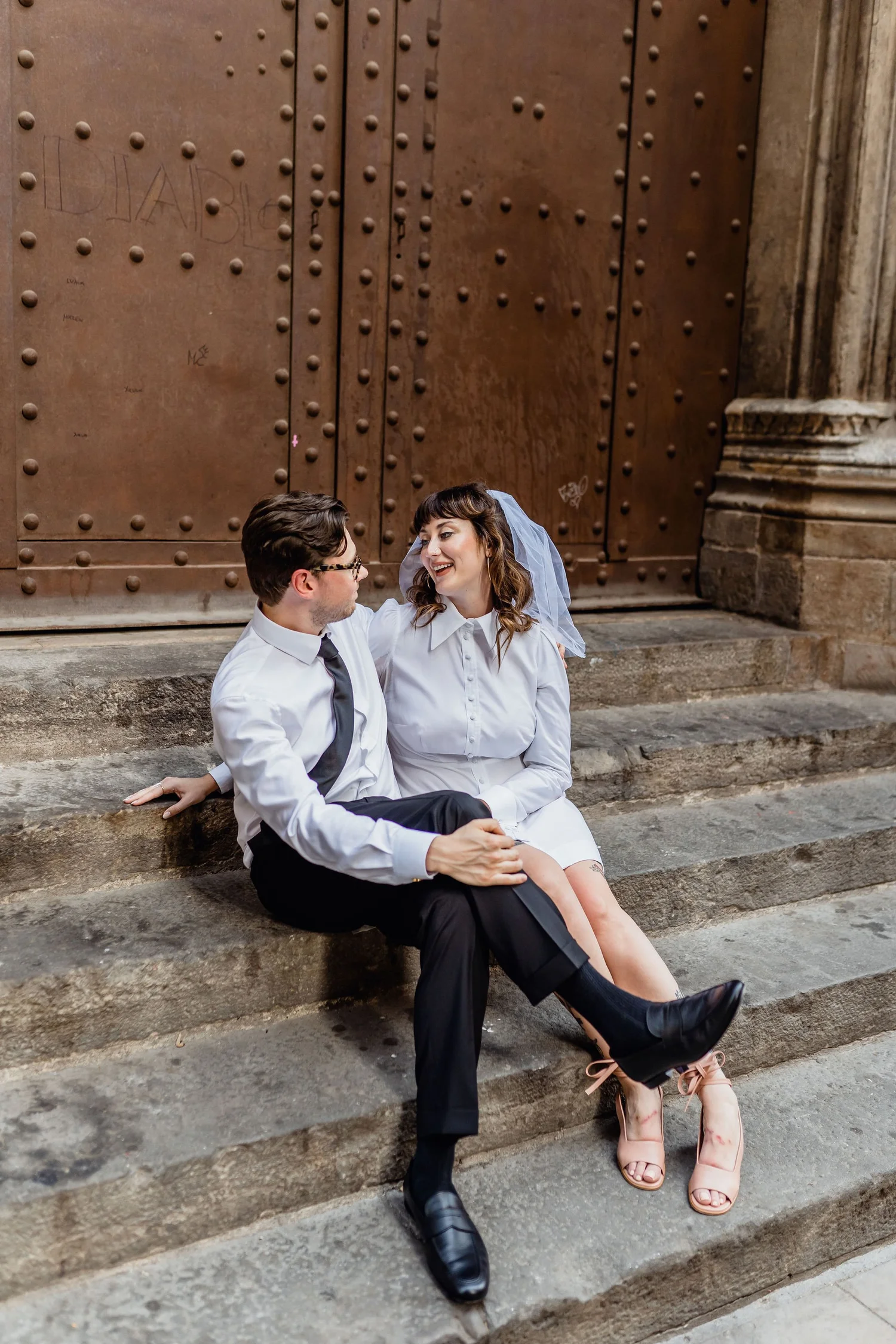 A couple dressed in white shirt and black pants sitting on stone steps, smiling and looking at each other in front of a large, brown door with metal studs.