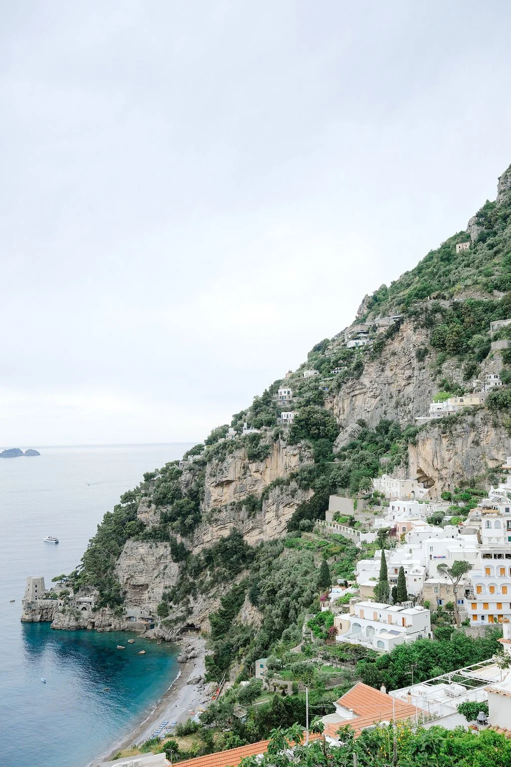 Steep hillside with white buildings and greenery overlooking the sea, with boats near the shore.