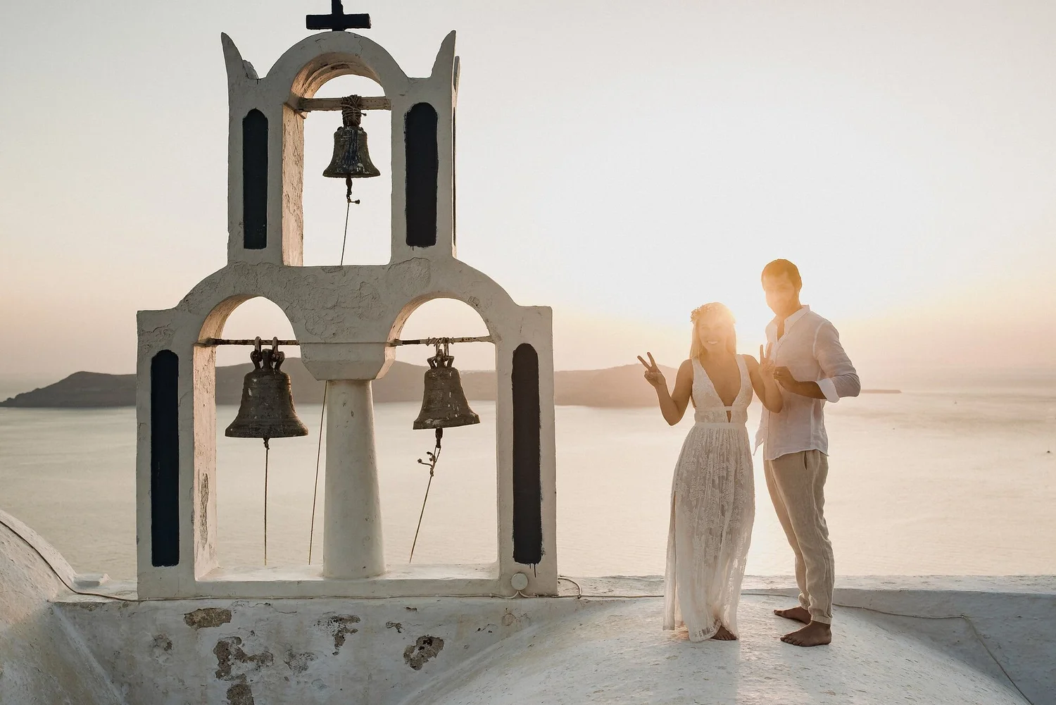 A couple standing on a beach at sunset, smiling and making peace signs, next to a white bell tower with multiple bells, overlooking the water and distant islands.