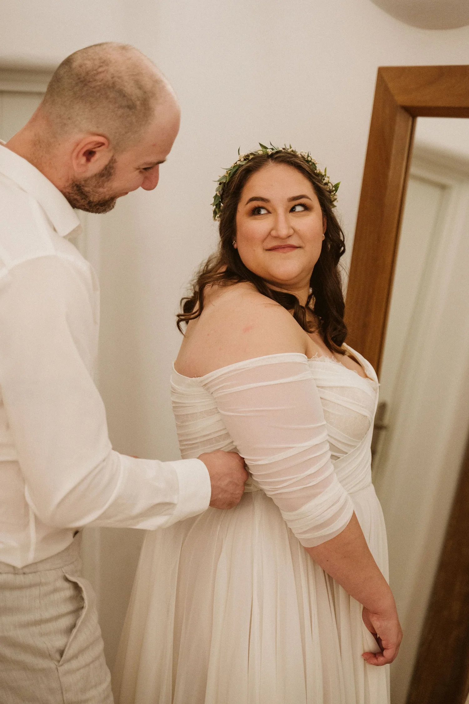 A bride and groom at their wedding, with the groom zipping up or adjusting the bride's wedding dress. The bride is wearing a white dress with off-the-shoulder sleeves and a floral crown, looking at the groom with a smile. The groom is dressed in a li