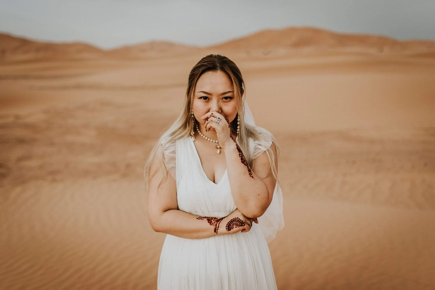 A woman with long hair and jewelry, wearing a white dress, standing in a vast desert landscape with mountains in the background.