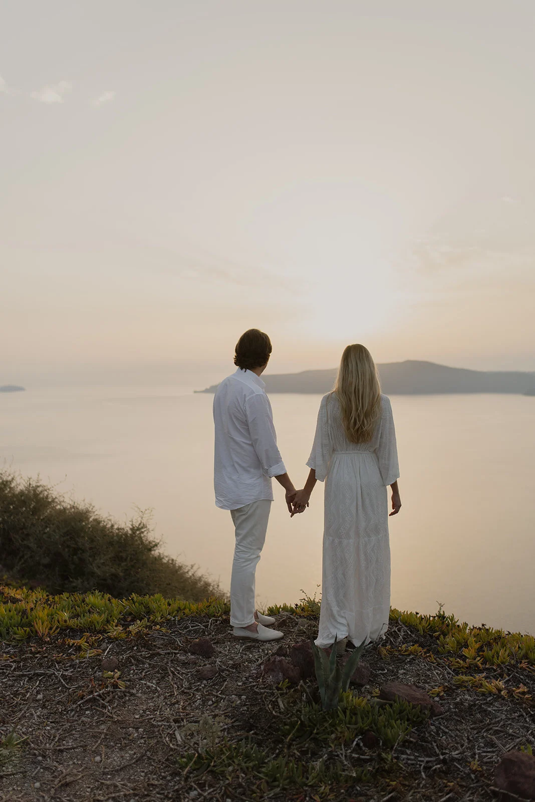 A couple holding hands and facing sunset overlooking water and distant hills, dressed in white, standing on a rocky and grassy terrain.