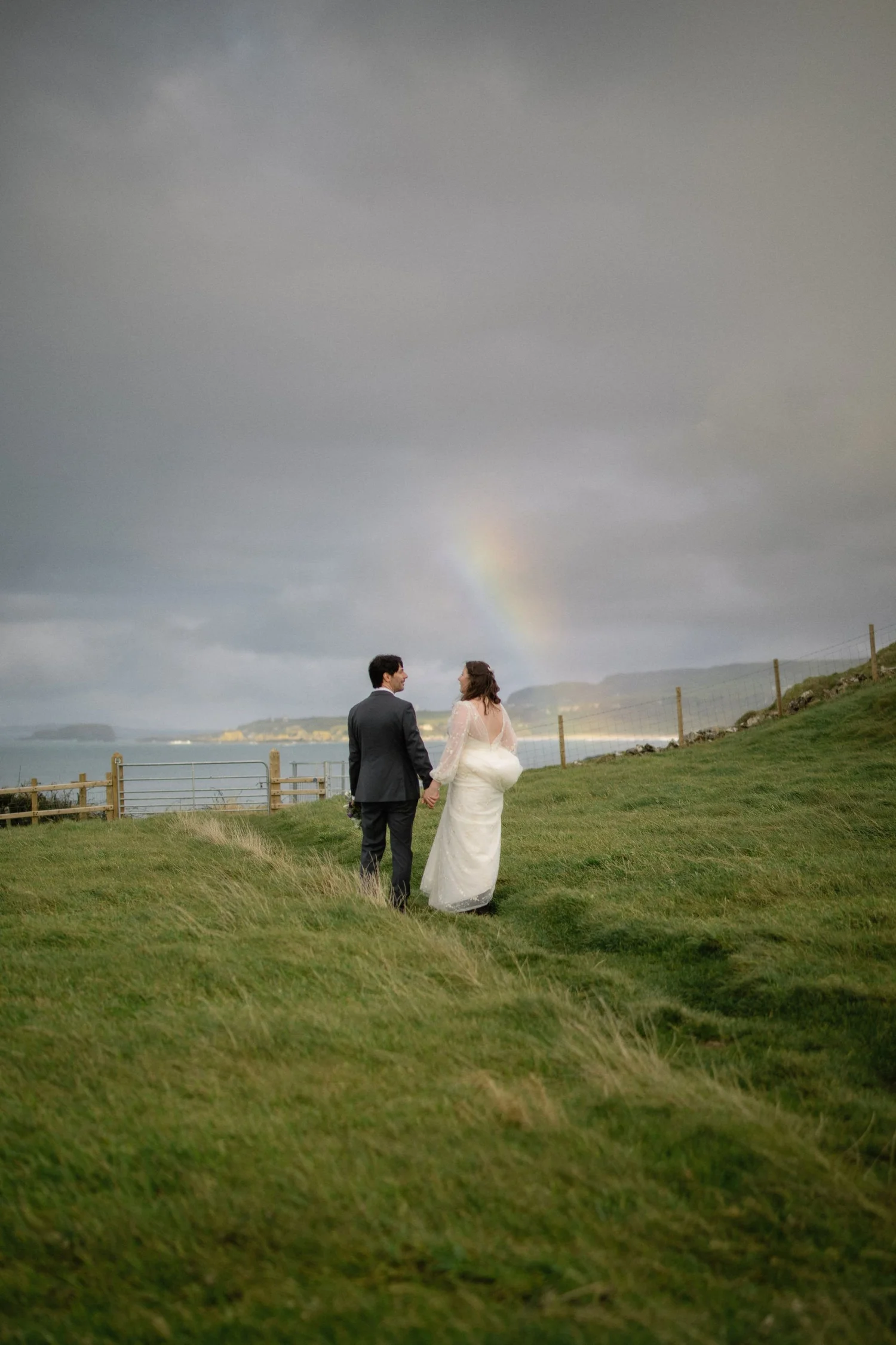 A bride and groom holding hands, standing on a grassy hill with a fence, overlooking the ocean, with grey stormy clouds and a faint rainbow in the sky.