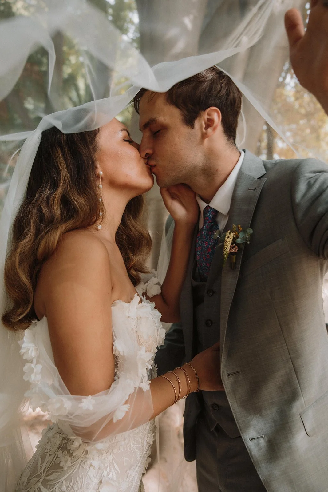 A bride and groom sharing a kiss under a sheer veil during their wedding, with trees visible in the background.