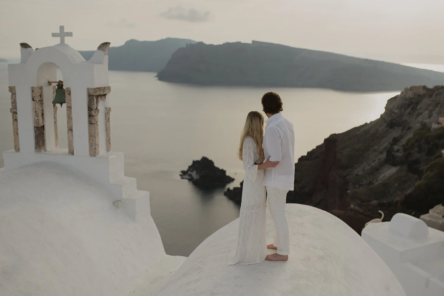 A romantic couple dressed in white clothing stands on a white rooftop overlooking the sea with islands in the background. The scene is calm and scenic, with soft natural lighting.