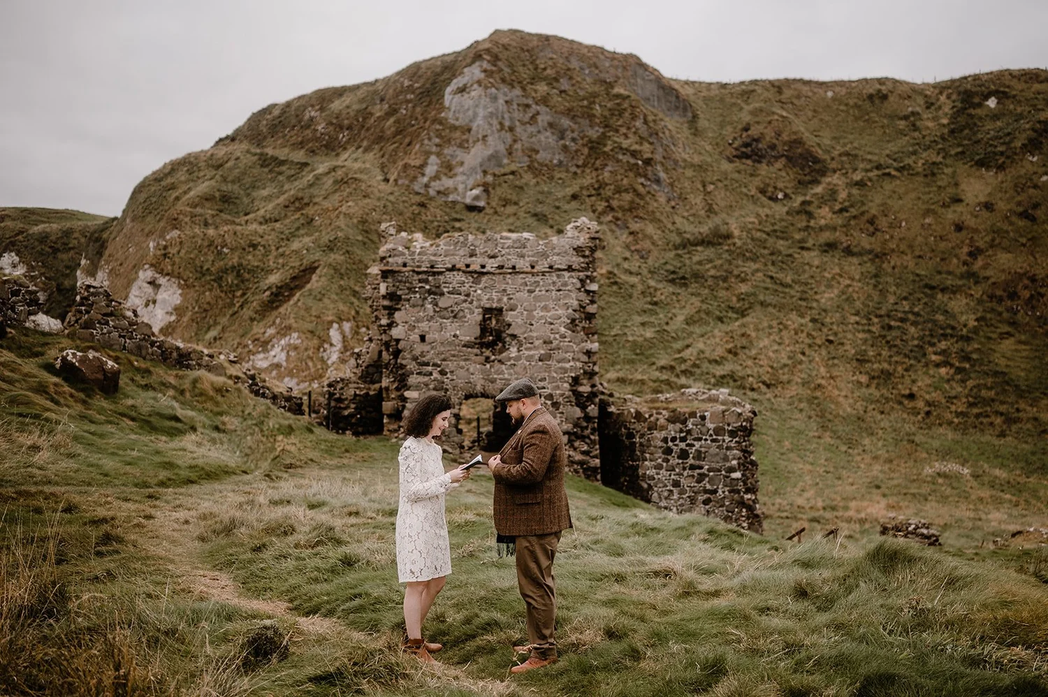 A couple standing outdoors on grassy terrain, exchanging vows or rings, with an ancient stone structure and rolling hills in the background.