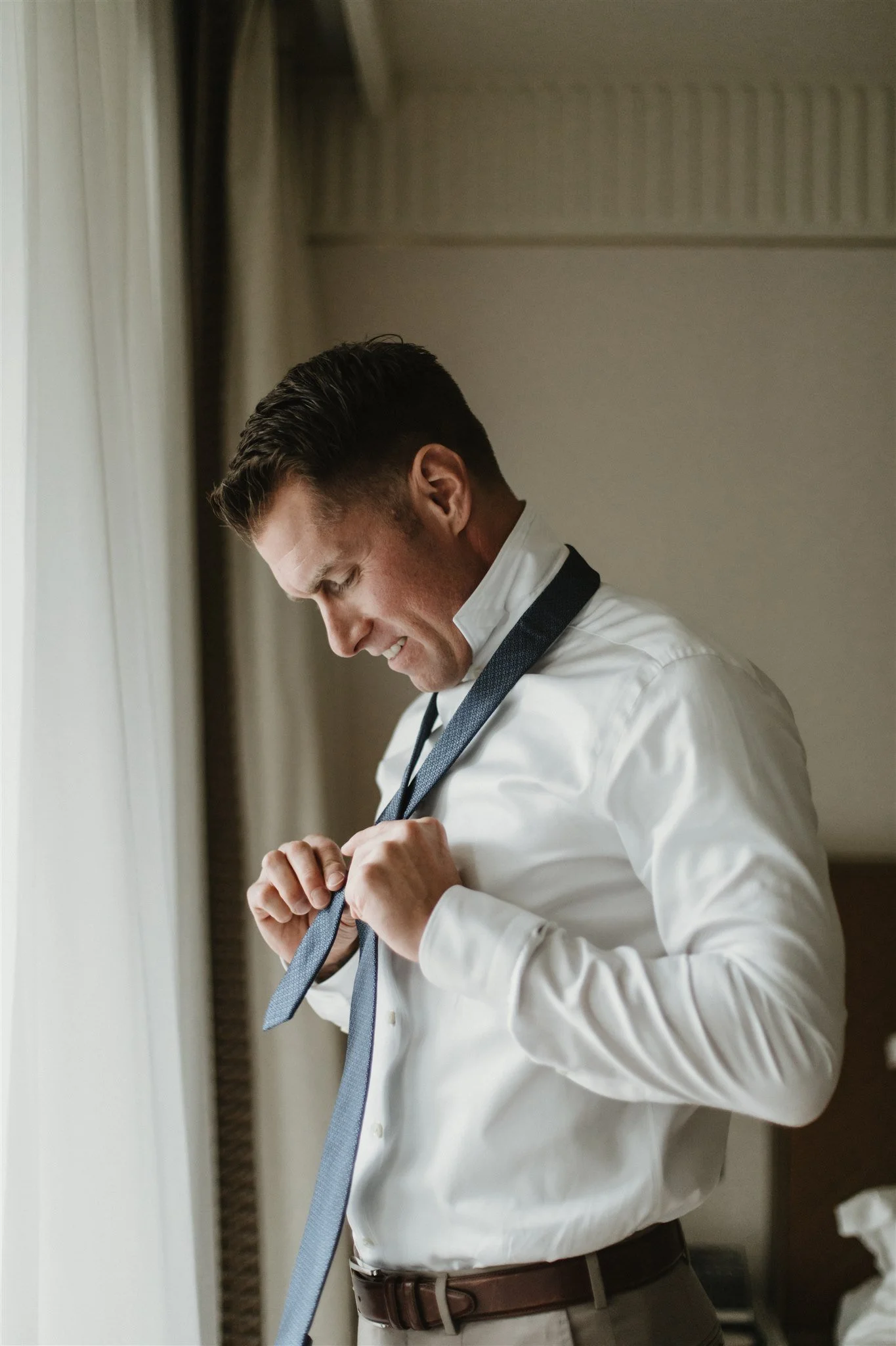 A man in a white shirt adjusting a blue tie near a window with curtains.