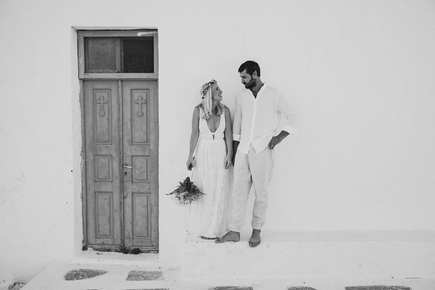 A black-and-white photo of a smiling woman in a white dress holding a bouquet, and a man in a white shirt and pants, standing against a plain white wall next to a weathered wooden door.
