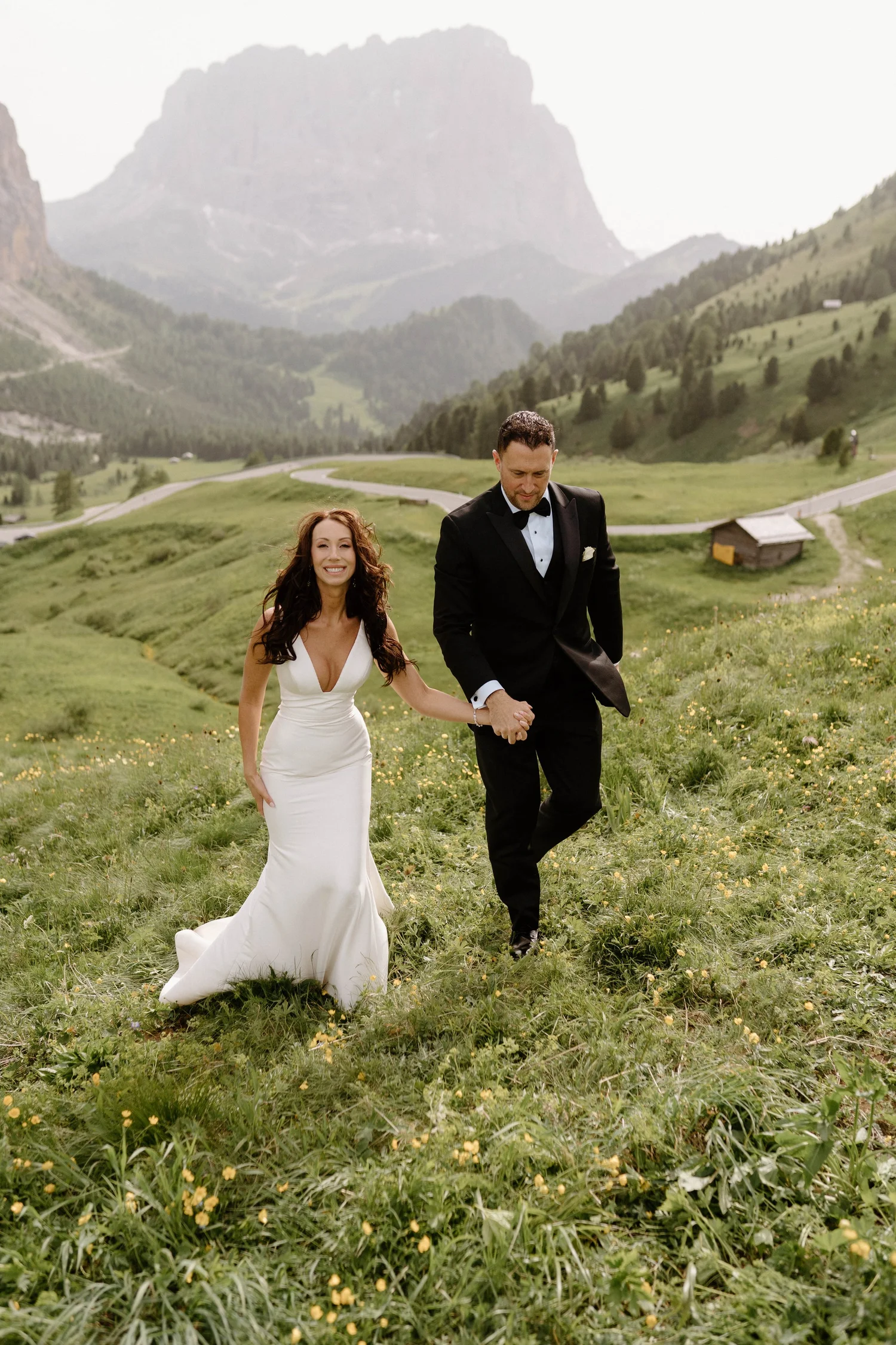 A bride and groom holding hands and running through a green meadow with mountains in the background.