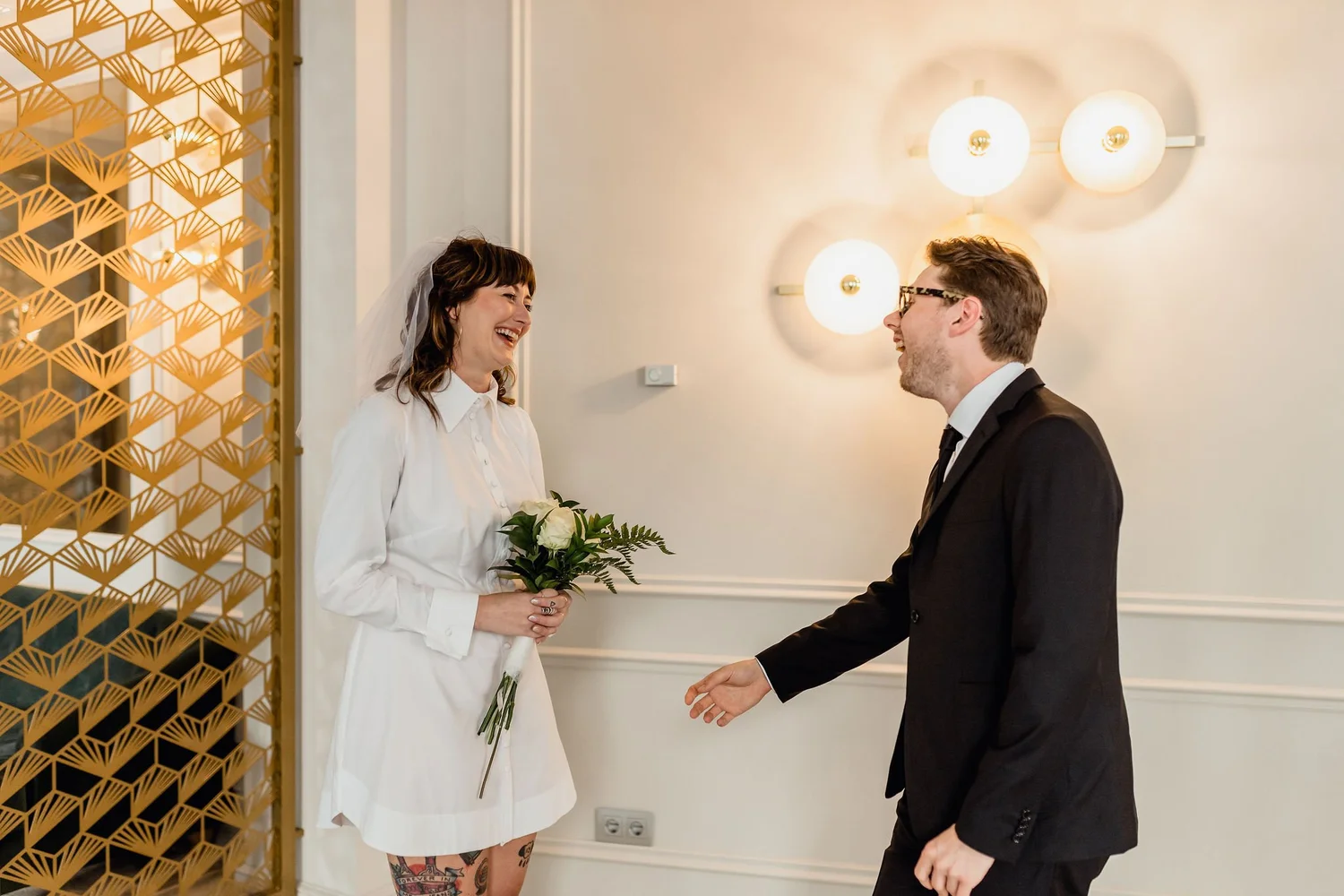A woman in a white shirt and veil holding a bouquet of white roses is smiling at a man in a black suit with glasses. They are standing indoors, near a decorative gold partition and modern lighting on the wall.
