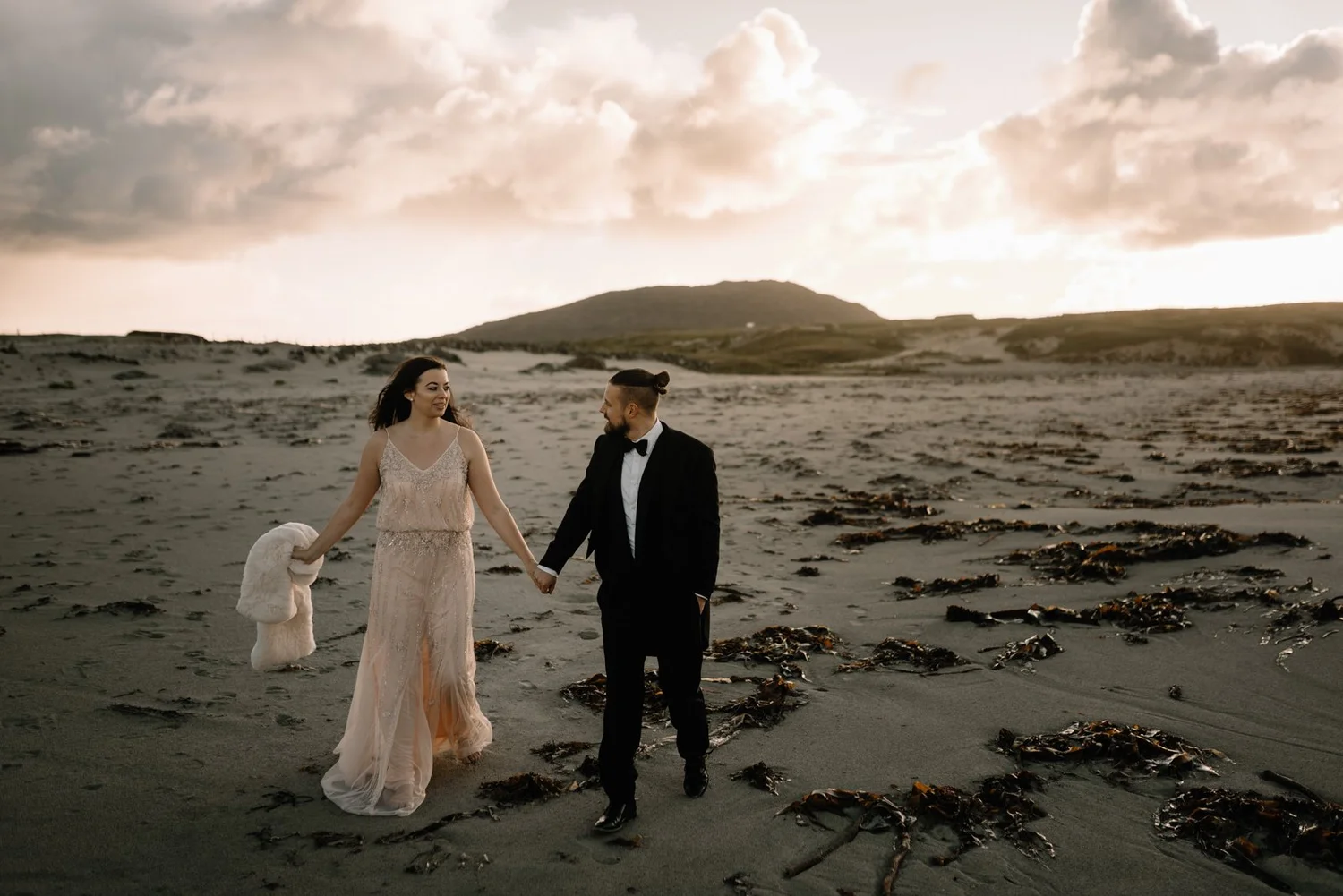A couple in wedding attire walking hand in hand on a beach at sunset, with seaweed scattered on the sand and a mountain in the background.