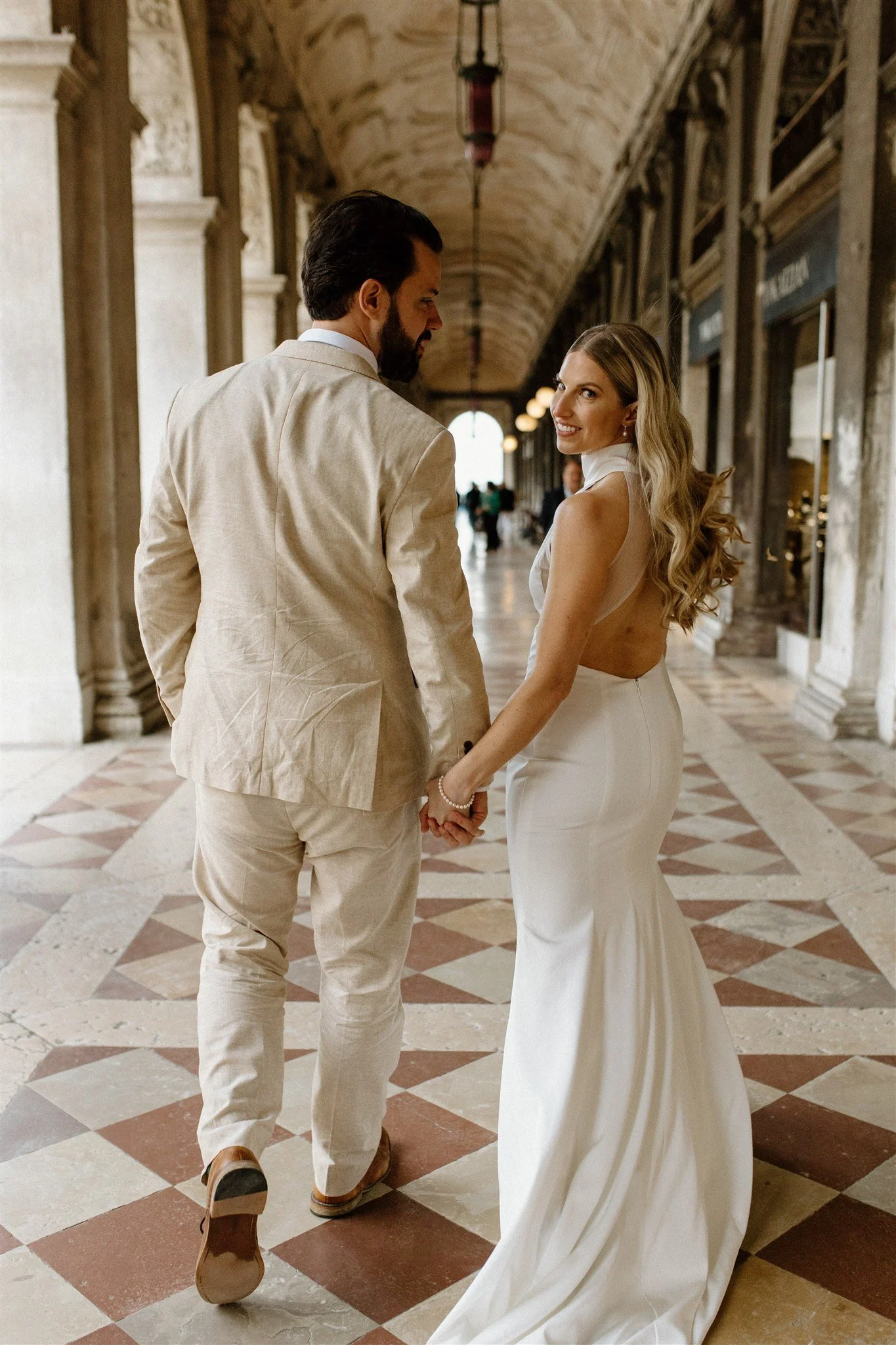 A couple holding hands and smiling at each other in a historic hallway with arched ceilings and patterned floors, dressed in wedding attire.
