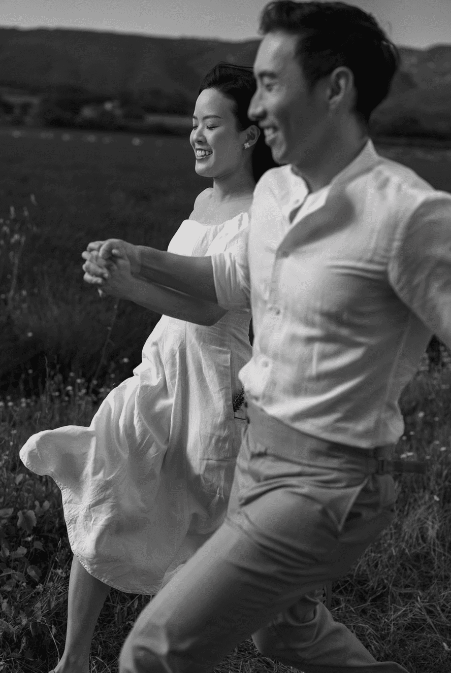 A black-and-white photo of a smiling couple sitting on grass outdoors, with rolling hills in the background, holding hands.