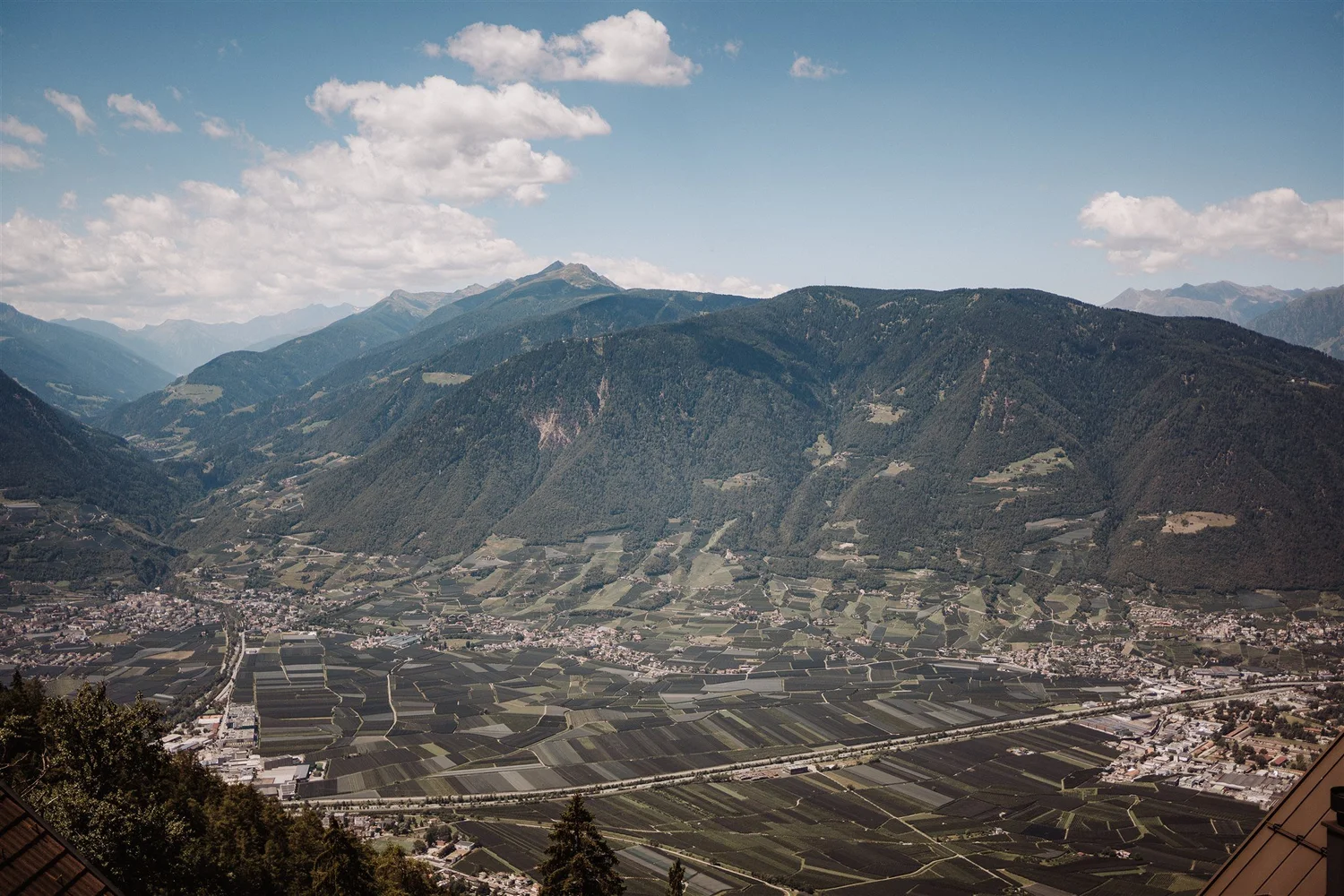 A scenic view of a valley with agricultural fields surrounded by lush green mountains under a partly cloudy sky.