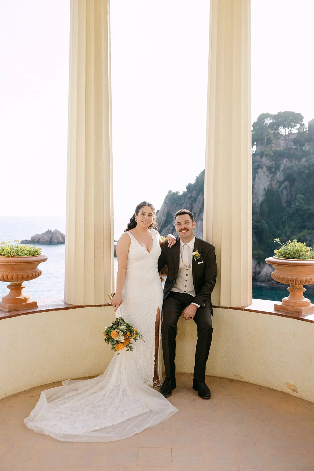 A newlywed couple, the bride in a white wedding dress holding a bouquet and the groom in a black suit, posing together on a balcony with ocean and rocky coastline scenery in the background.