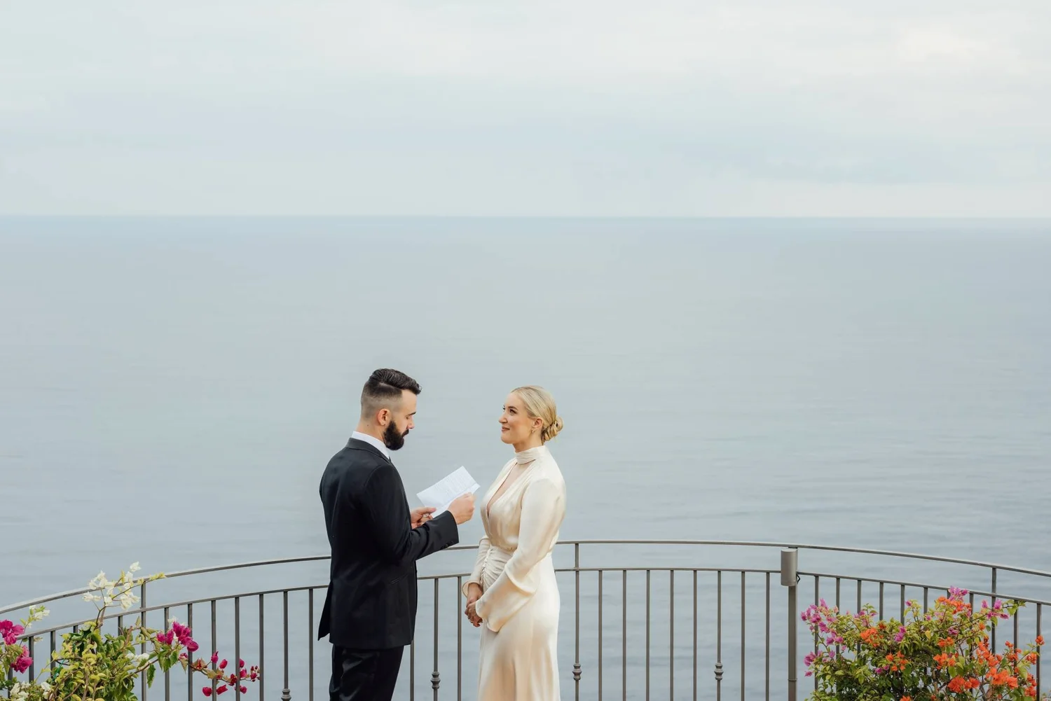 A man and woman in wedding attire exchange vows outdoors near a body of water, with colorful flowers along a metal railing.