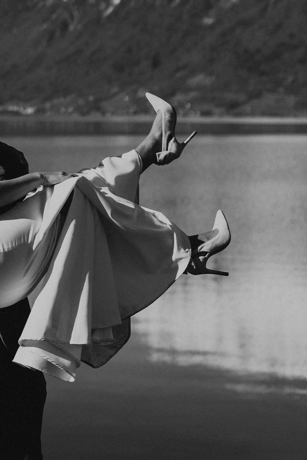 Black and white photo of a woman in a flowing dress and high heels sitting near a body of water with a mountain background, her legs extended and crossed at the ankles.