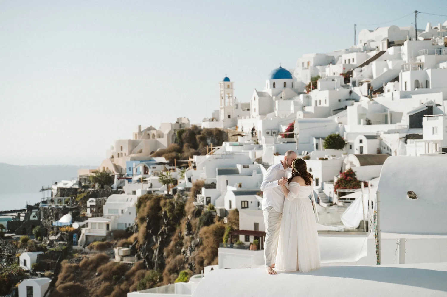 A couple dressed in white wedding attire standing outdoors on a white platform with a picturesque backdrop of white buildings with blue domes on a hillside in Santorini, Greece.
