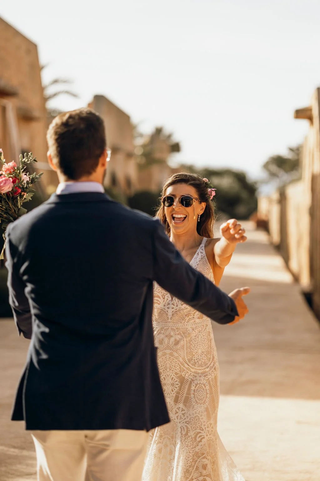 A couple on their wedding day, with the bride wearing sunglasses and smiling, embracing outdoors on a sunny day.