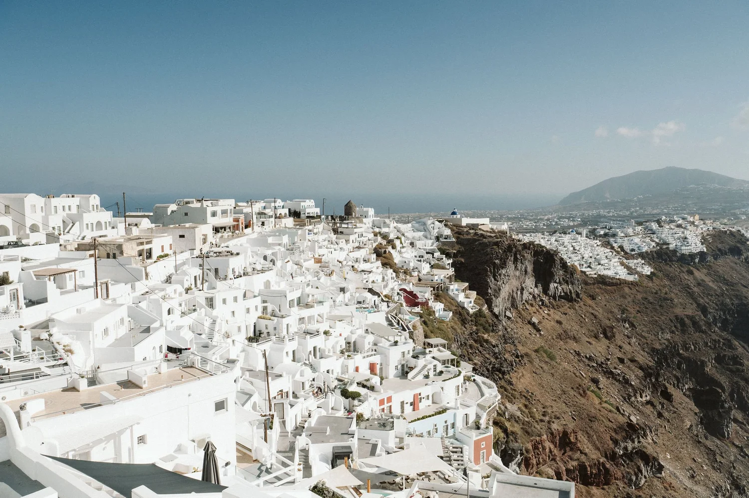 A scenic view of a hillside town with white buildings, possibly in Greece, overlooking the ocean and a distant mountain under a clear blue sky.