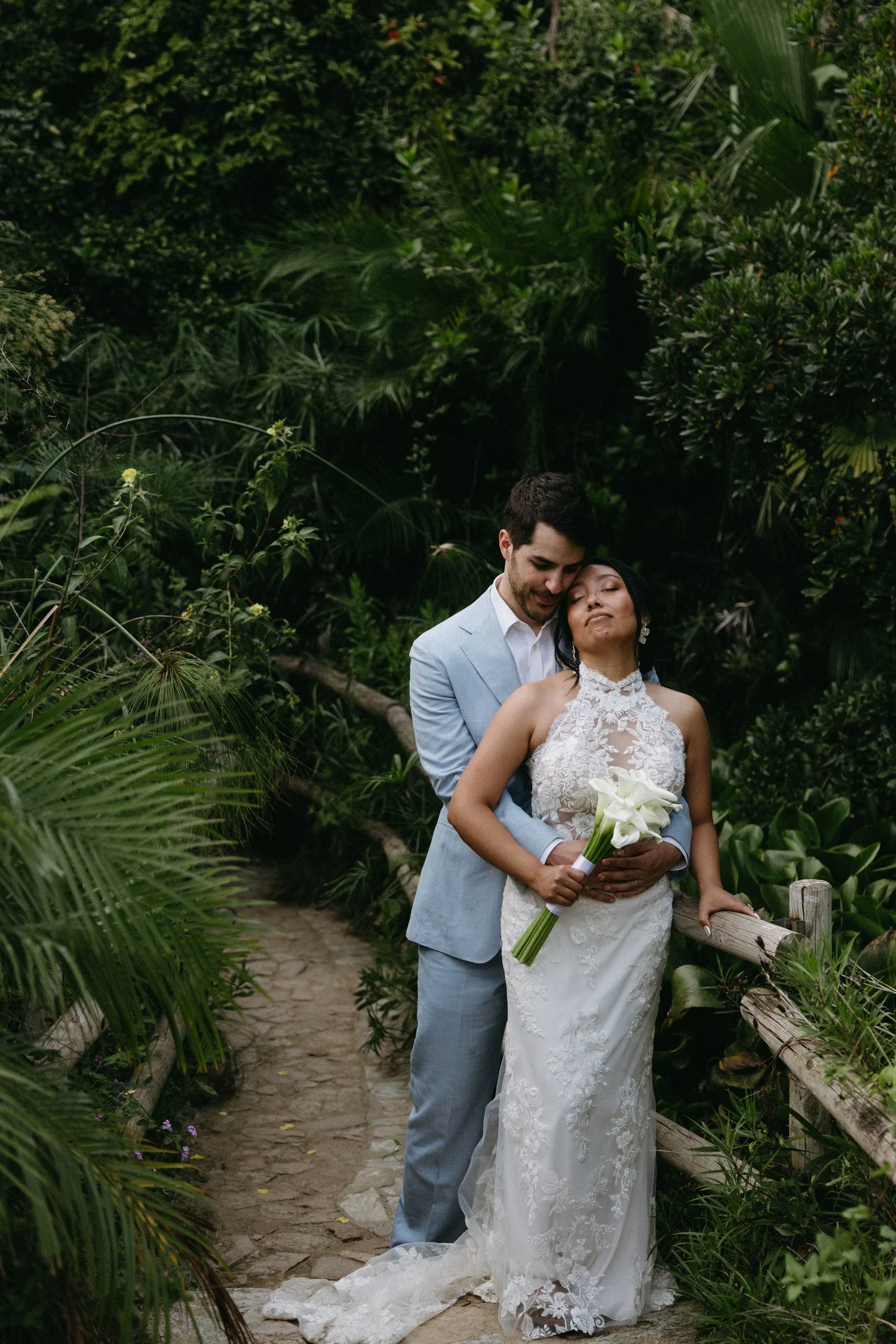 A newlywed couple in wedding attire standing on a garden pathway, surrounded by lush green foliage, with the bride holding a bouquet of white flowers and the groom hugging her from behind.