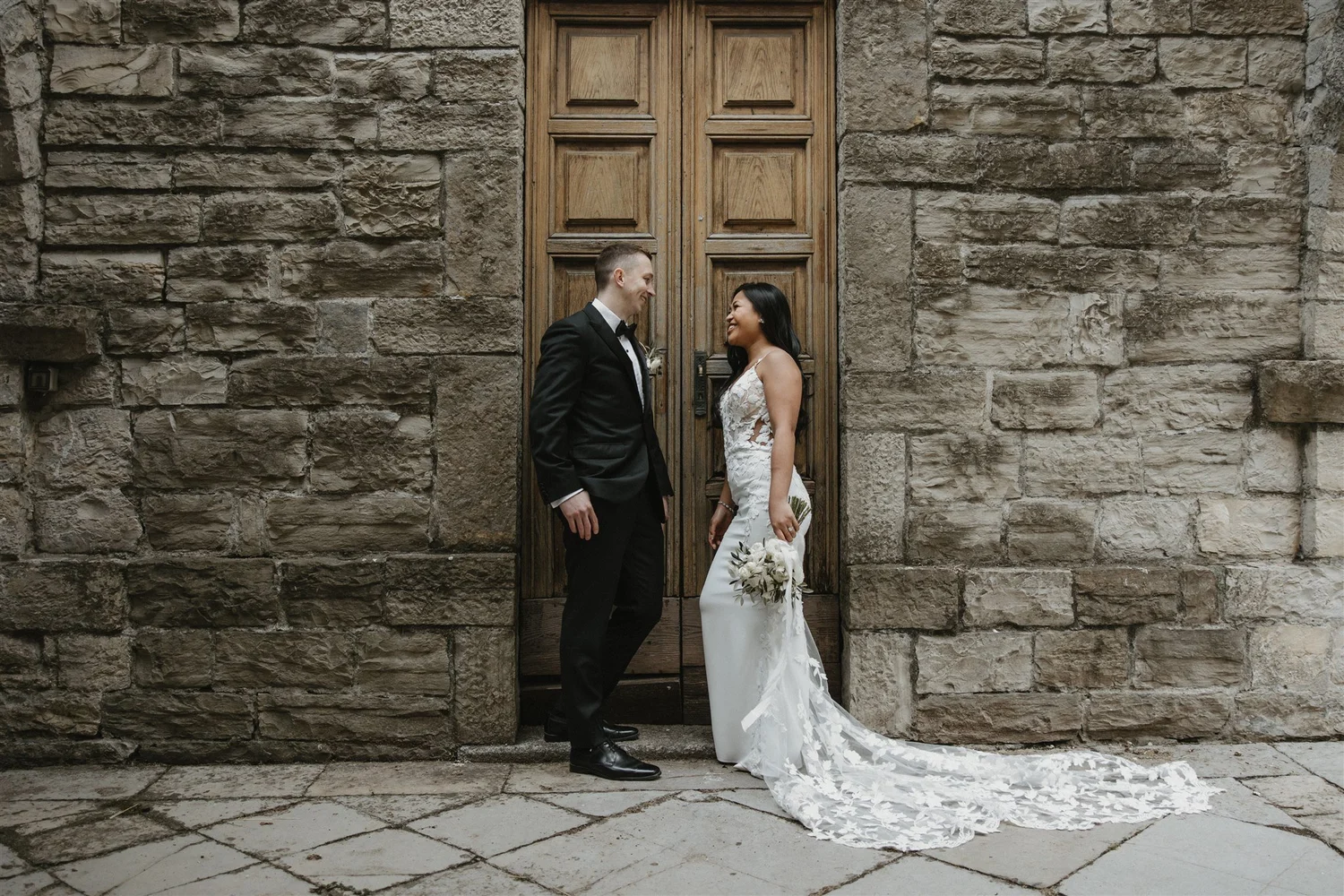 A bride and groom stand face to face in front of a large wooden door with stone walls on either side. The groom is in a black tuxedo with a bowtie, and the bride is in a white lace wedding dress holding a bouquet of flowers. They are smiling at each