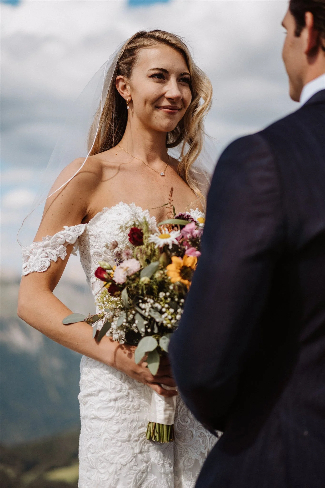 A bride and groom stand outdoors under a cloudy sky, facing each other during their wedding ceremony. The bride wears a lace wedding dress and holds a colorful bouquet of flowers. The groom is dressed in a dark suit, and they express happiness as the