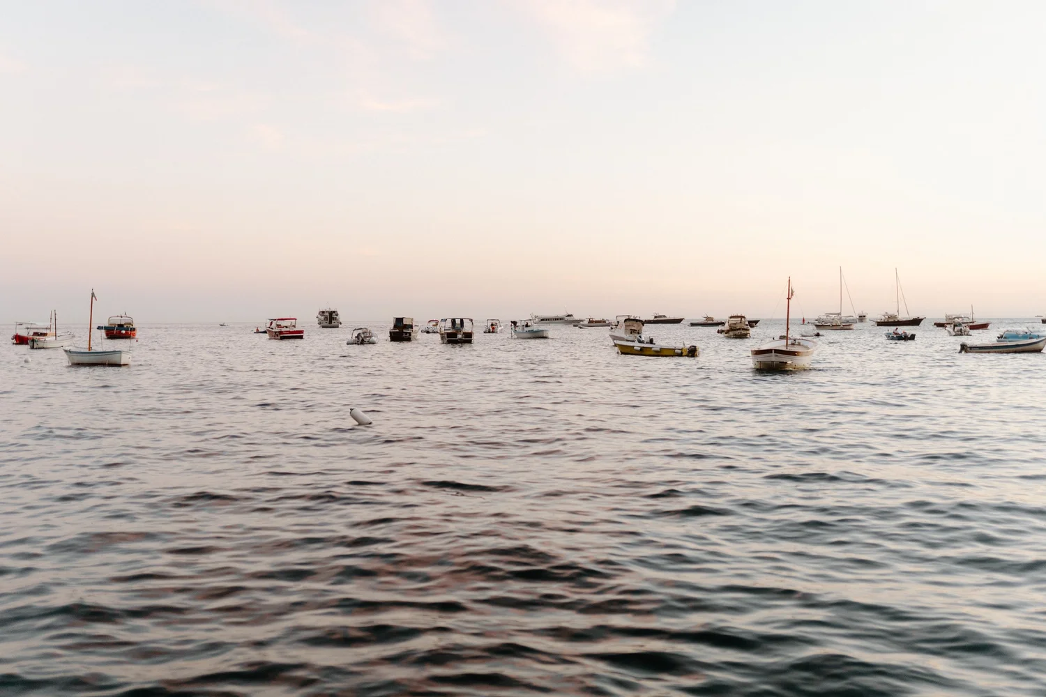 A serene scene of a harbor with numerous boats floating on calm water during sunset or sunrise, under a lightly clouded sky.
